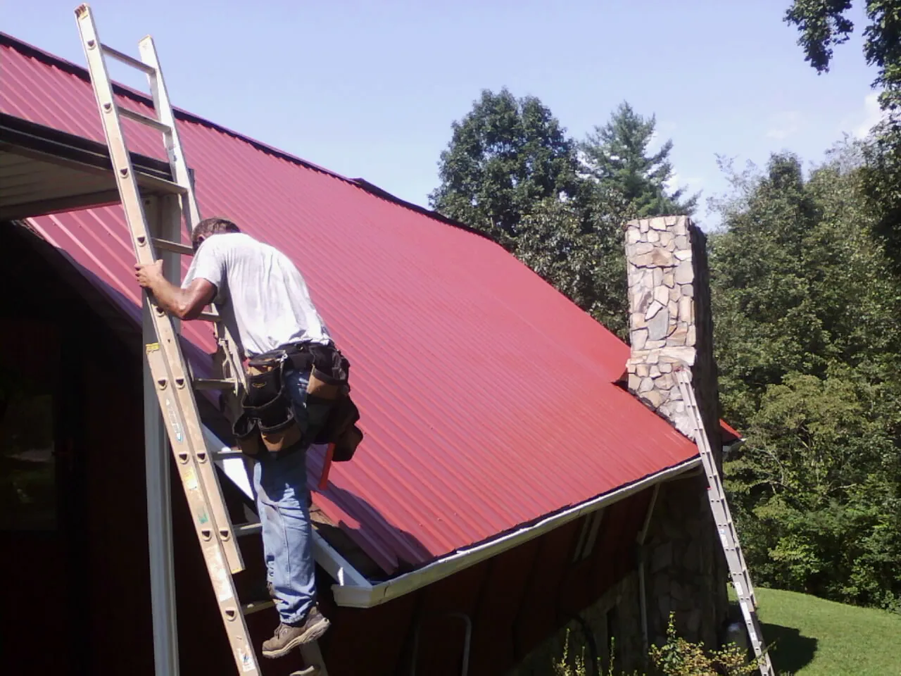 Roofer on a ladder beside a red metal roof and stone chimney — Soffit & Fascia Repair by Roofing Brothers in Moultrie